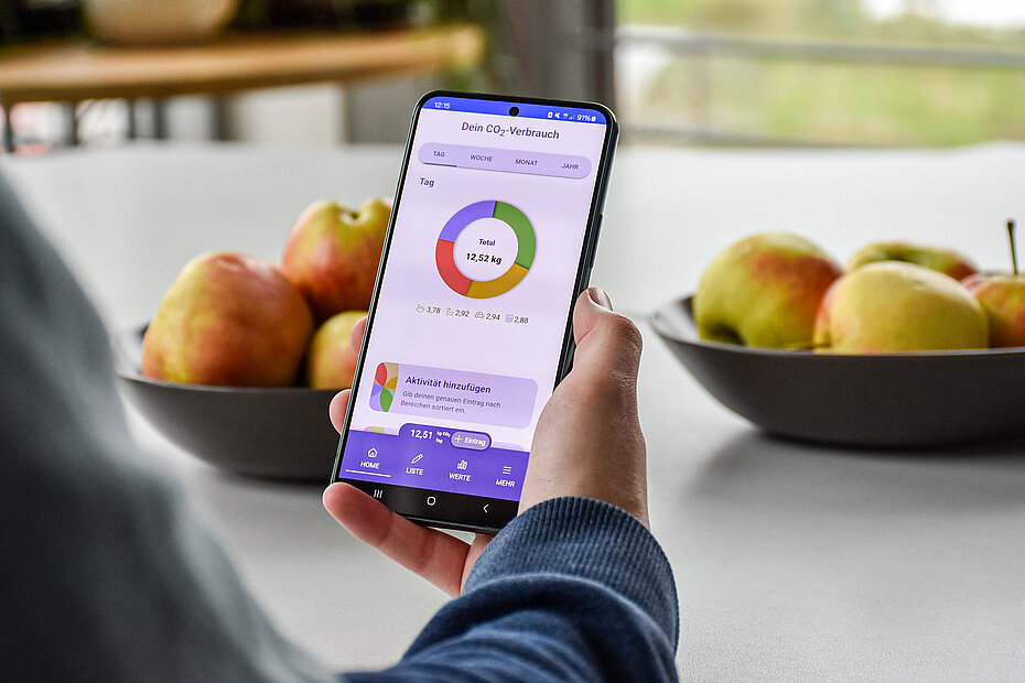 Eine Person hält ein Smartphone in der Hand. Auf dem Bilschirm ist die App "CO2-Diary" geöffnet. Im Hintergrund stehen zwei Schalen mit Äpfeln.