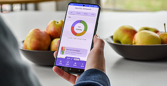 Eine Person hält ein Smartphone in der Hand. Auf dem Bilschirm ist die App "CO2-Diary" geöffnet. Im Hintergrund stehen zwei Schalen mit Äpfeln.